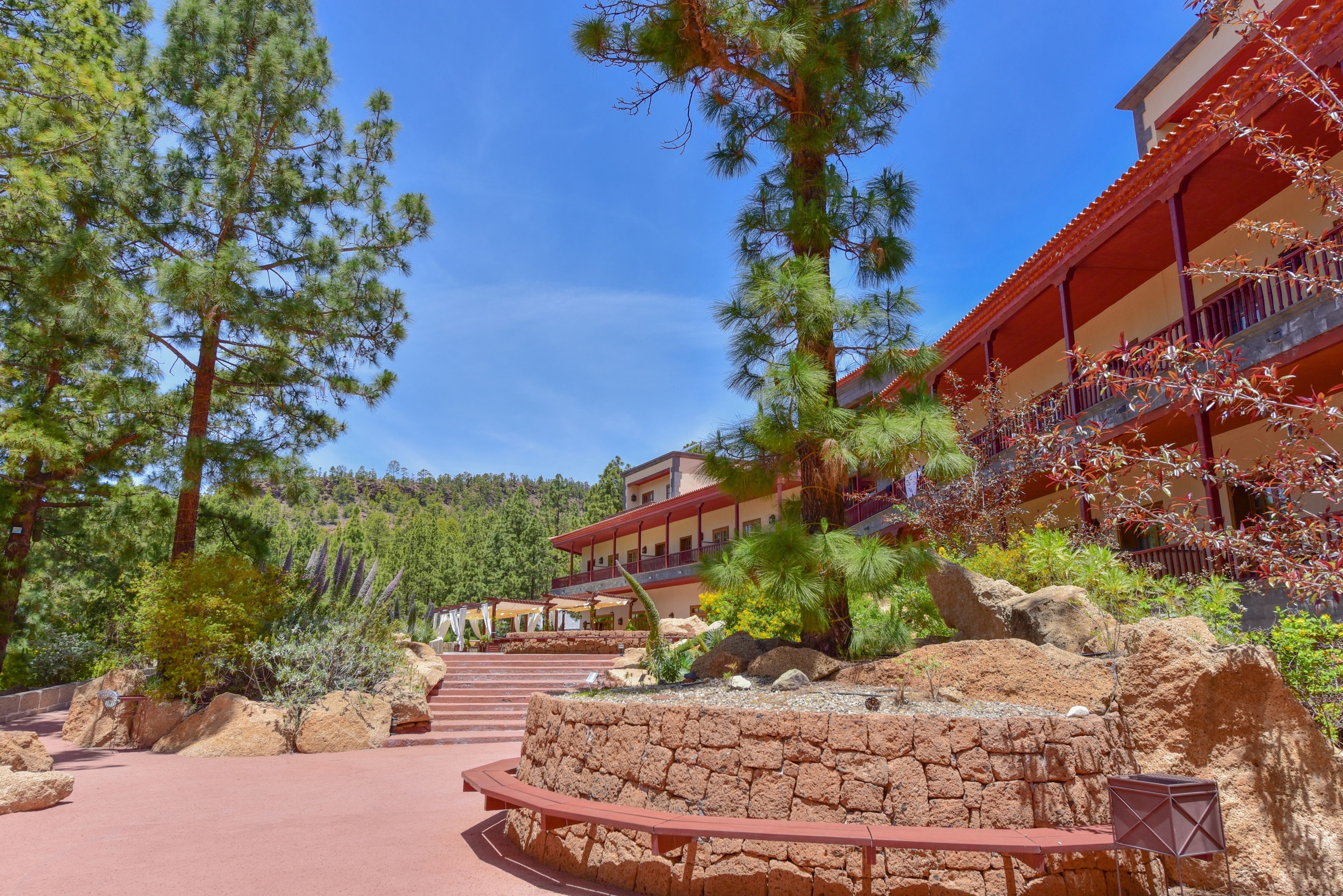 A resort with a stone pathway leading to a building with red and beige exterior walls, surrounded by pine trees and a mountainous backdrop.