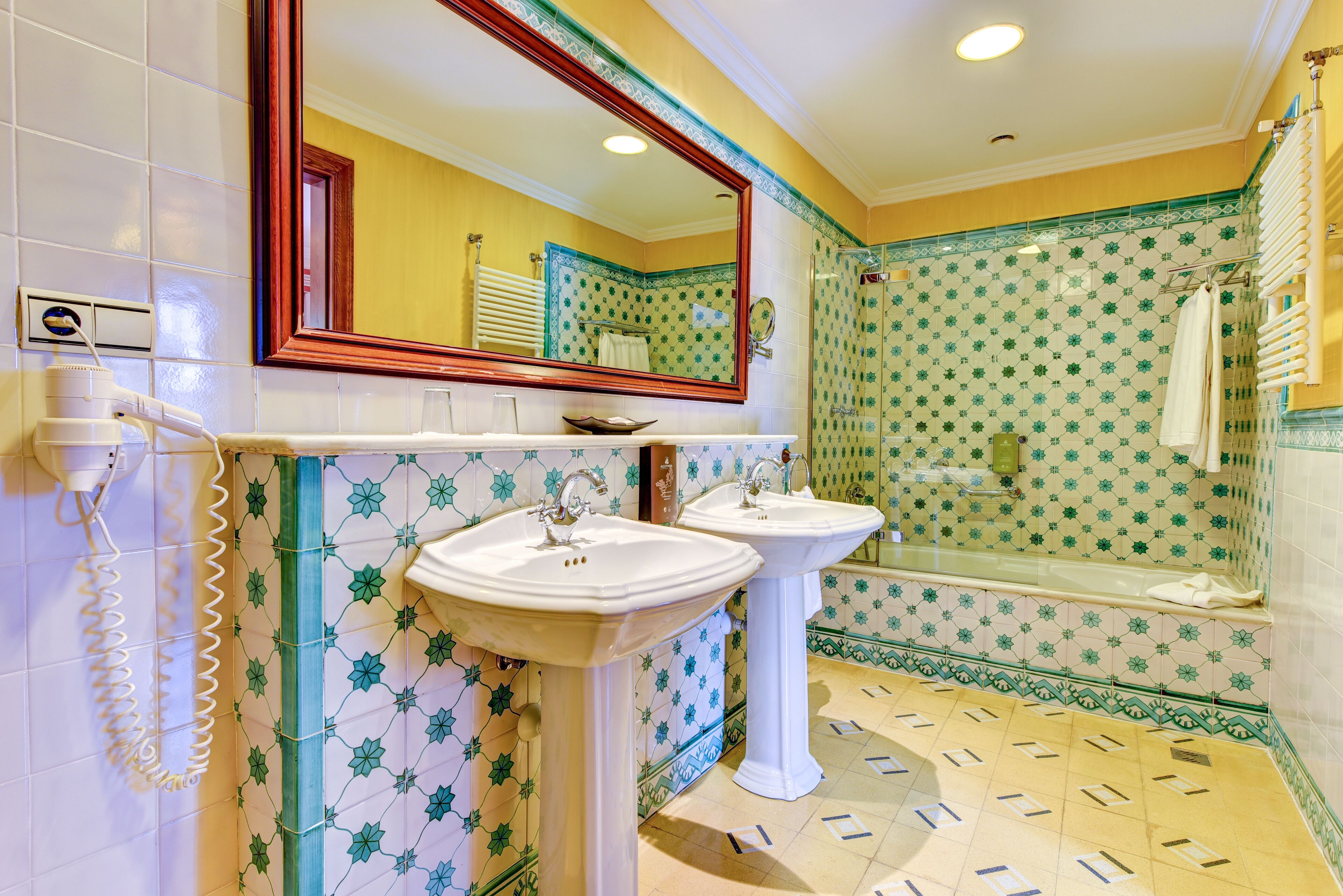 Un baño con dos lavamanos blancos, un espejo grande y una zona de ducha con azulejos estampados.
