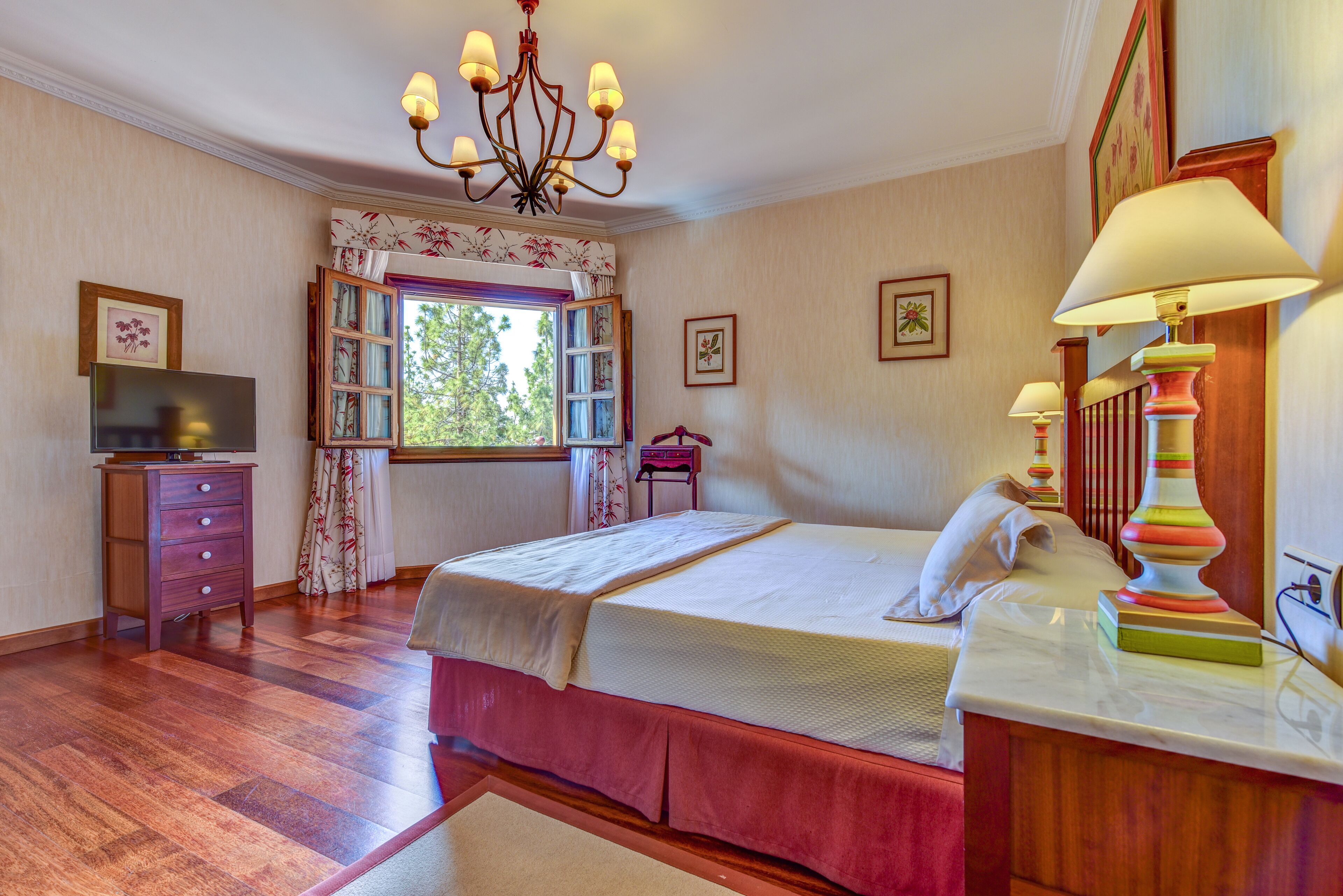 A bedroom with a bed, dresser, TV, window with curtains, and a chandelier.