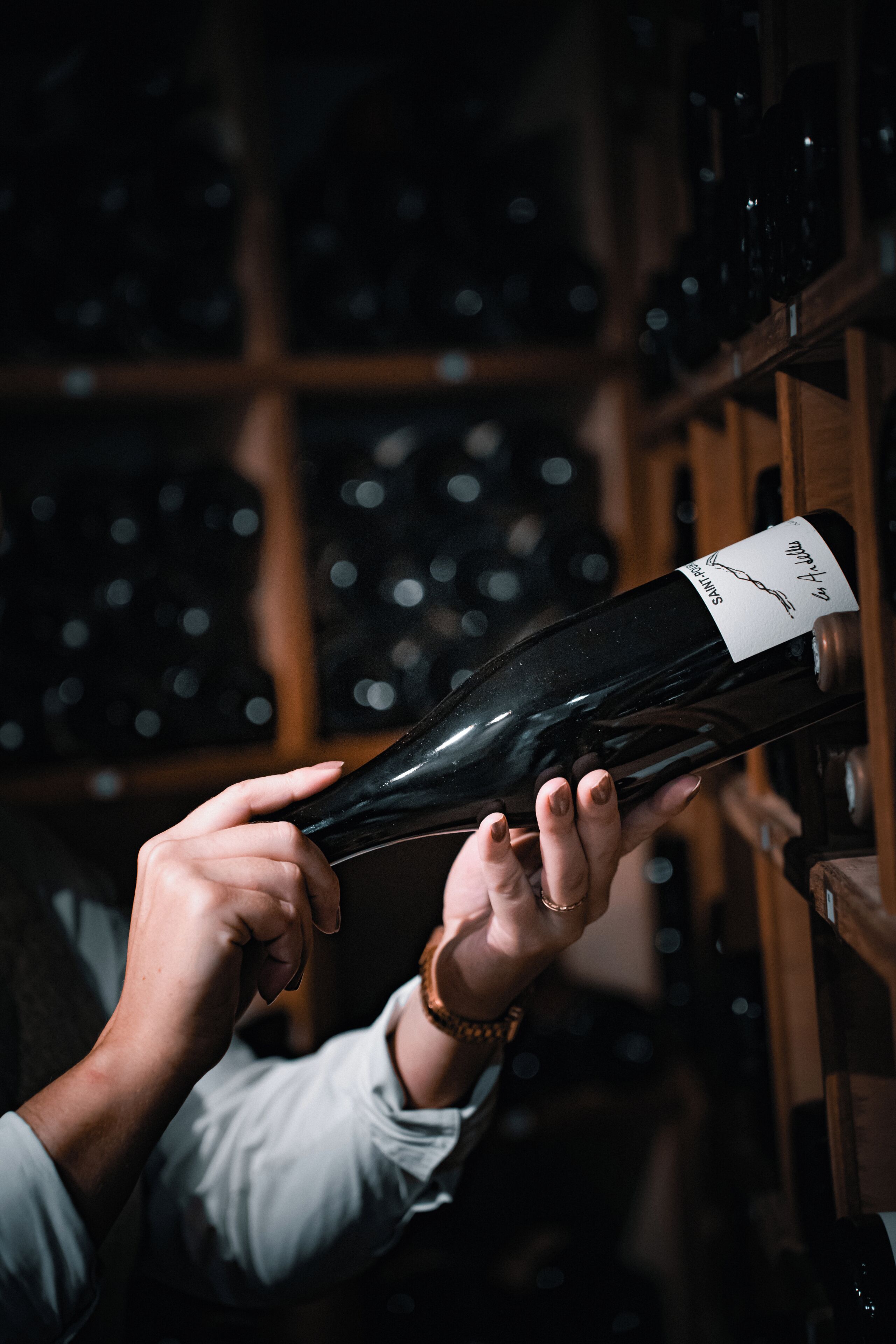 Uma pessoa segurando uma garrafa de vinho com etiqueta, em um quarto com pouca luz e prateleiras cheias de garrafas de vinho ao fundo.