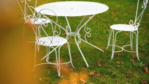 Ein Tisch mit Stühlen aus weißem Metall steht auf einer grünen Rasenfläche, im Hintergrund ist eine Steinmauer zu sehen.
