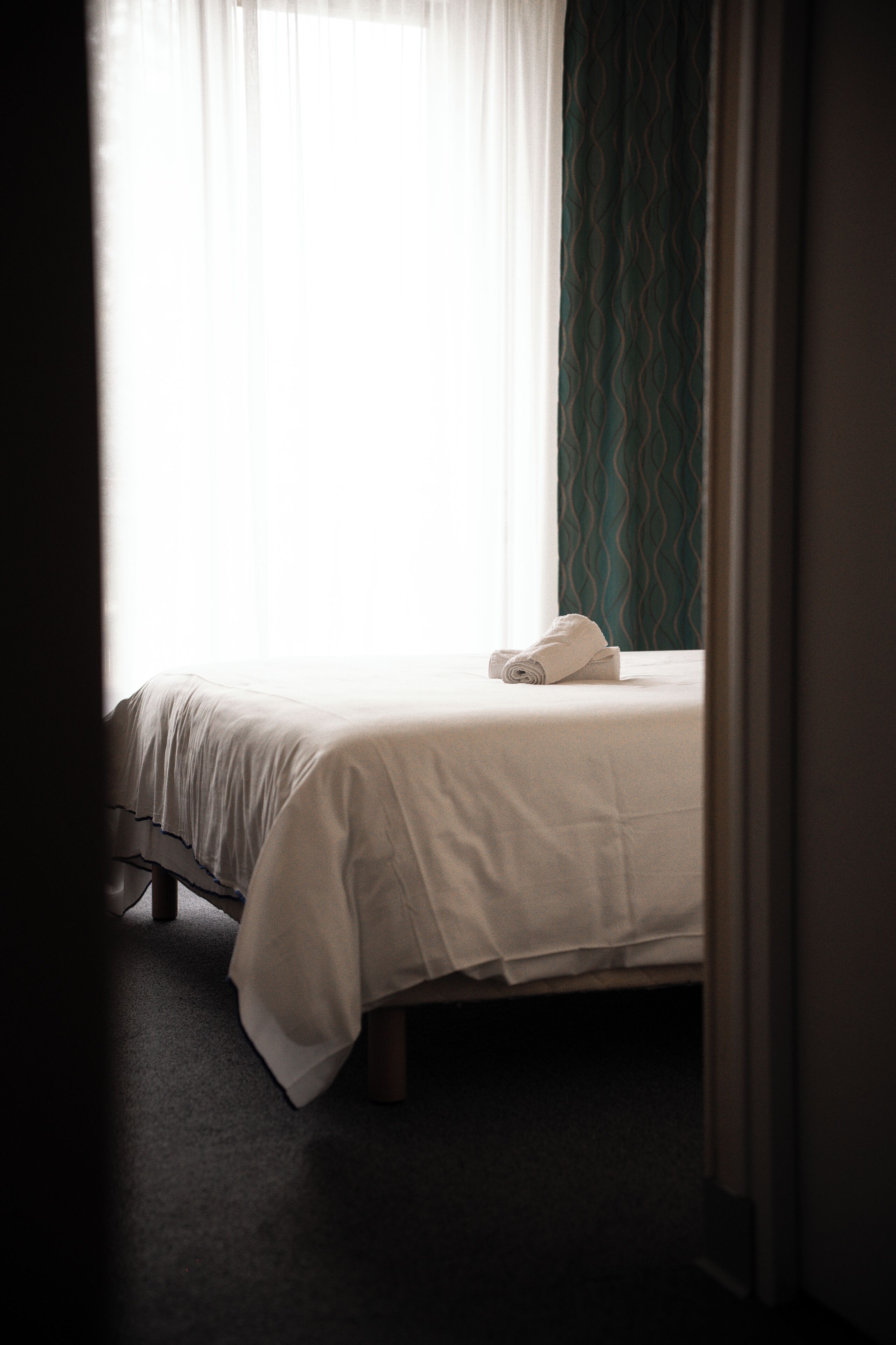 A hotel room with a bed, a rolled towel, and a window with curtains.