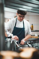 Um cozinheiro com camisa e avental branco prepara comida em uma cozinha com eletrodomésticos de aço inoxidável e painel de azulejos.