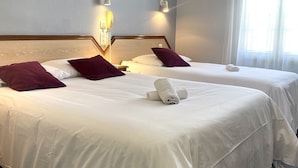 Ein Hotelzimmer mit zwei Betten, jeweils mit weißer Bettwäsche und violetten Kissen. An der Wand befinden sich Wandlampen und ein Telefon.