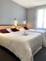 Quarto de hotel com duas camas, cada uma com roupa de cama branca e almofadas roxas. Há luminárias fixadas na parede e um telefone na parede.