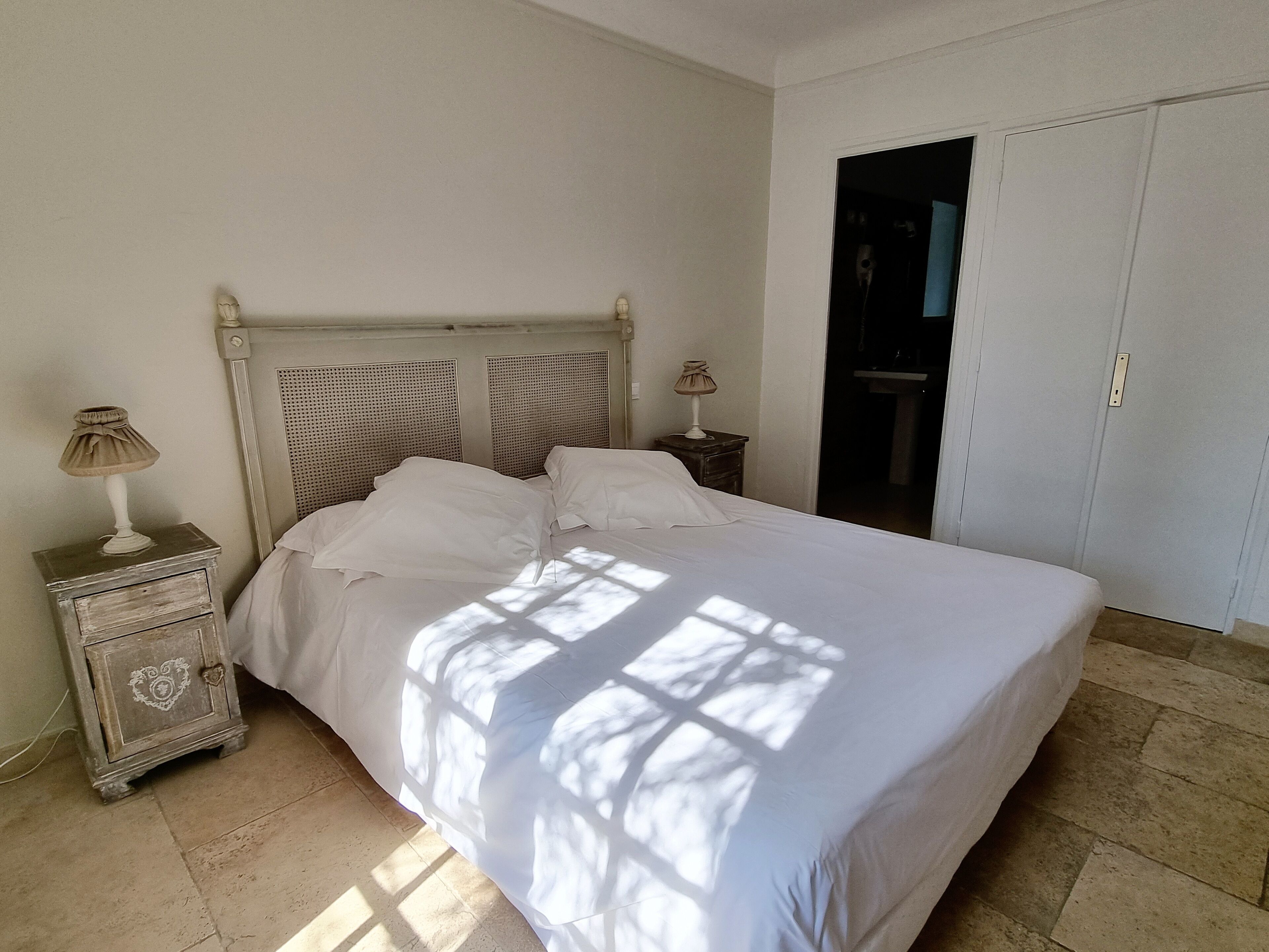A neatly made bed with white linens in a room with a classic design, including a wooden nightstand with a lamp and a dresser with a heart-shaped design.