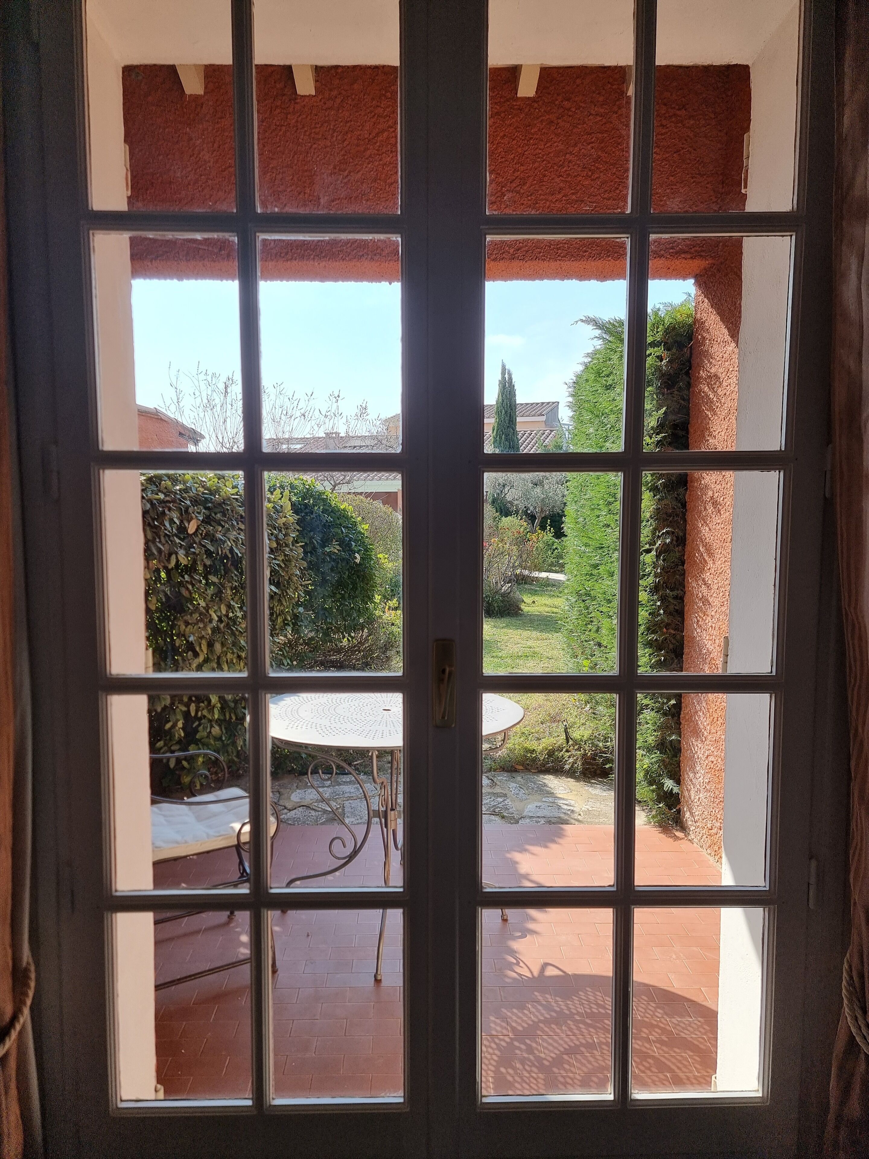 A view through a window with a view of a garden, a white table, and a brick floor.