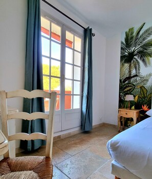 A room with a wooden chair, a bed, a window with curtains, and a wall-mounted lamp. - Castillon des Baux (Maussane-les-Alpilles)