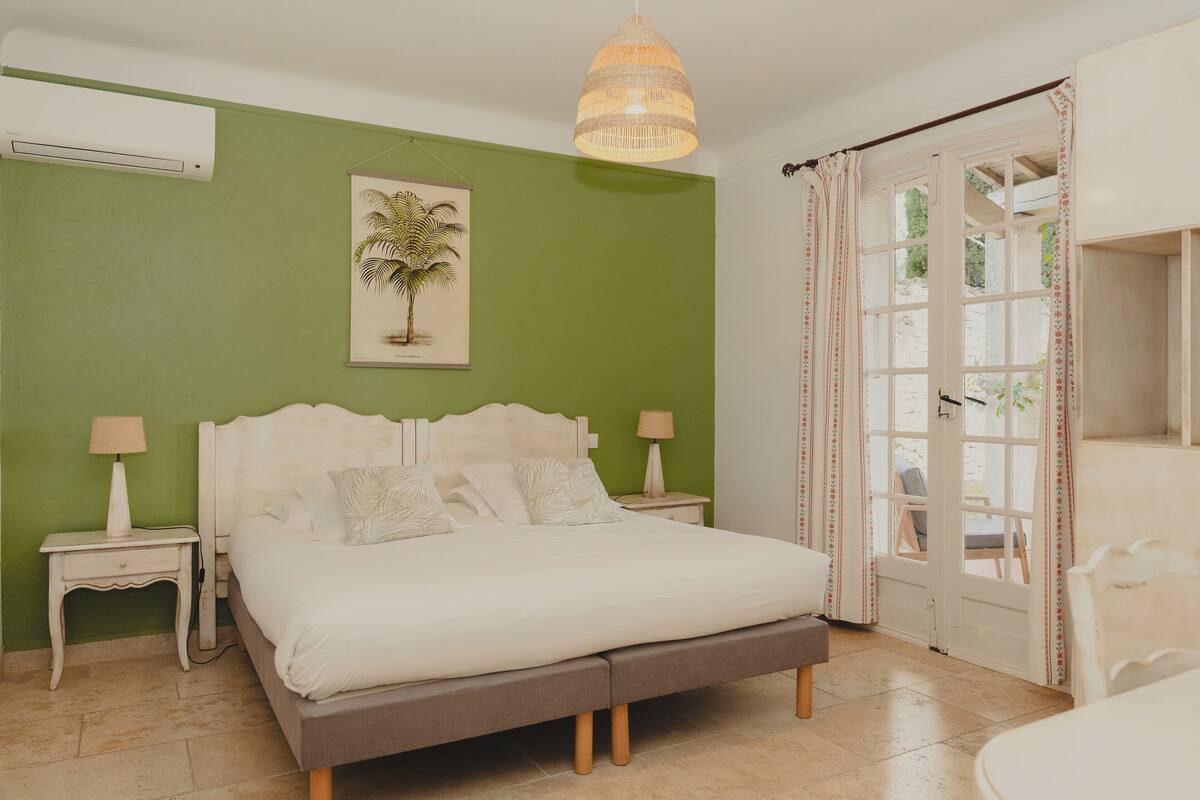 A bedroom with a bed, bedside tables, a hanging light fixture, a framed picture, and a view through a door.