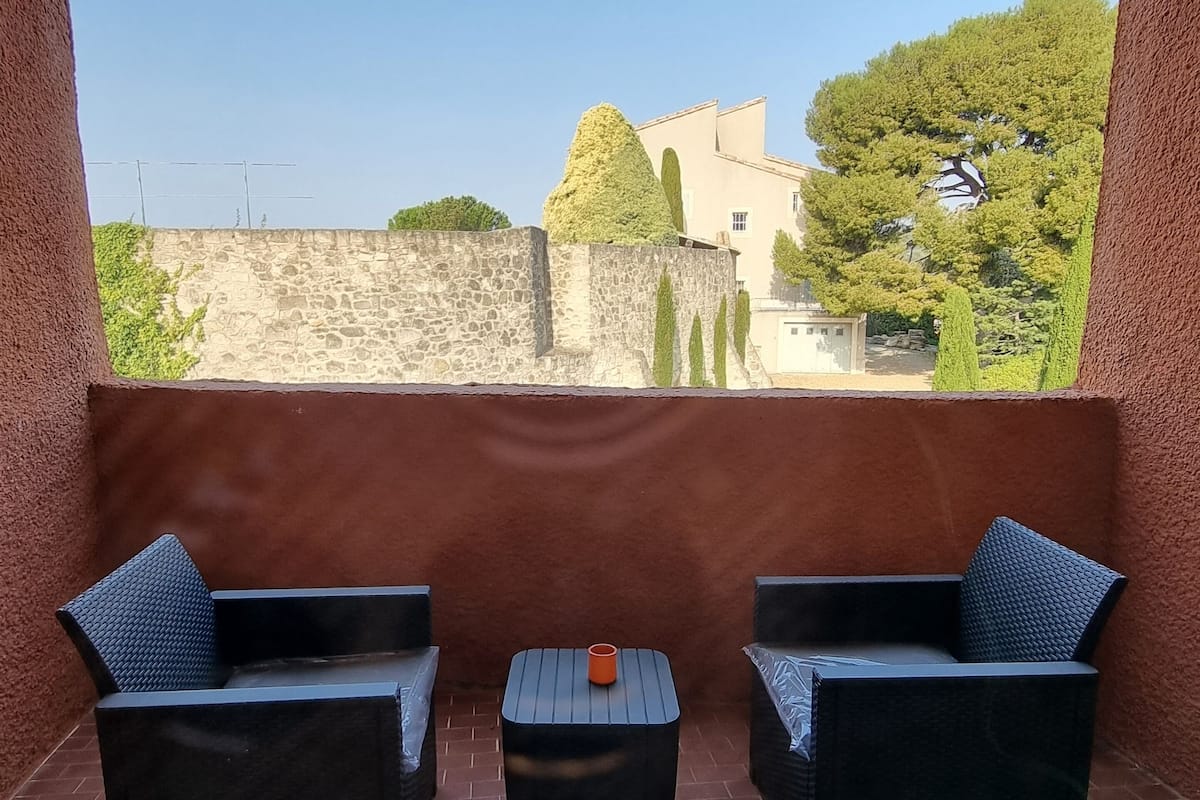 A balcony with two wicker chairs and a small table, a stone wall, and a view of buildings and trees.