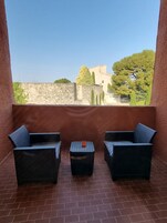 A balcony with two wicker chairs and a small table, a stone wall, and a view of buildings and trees.