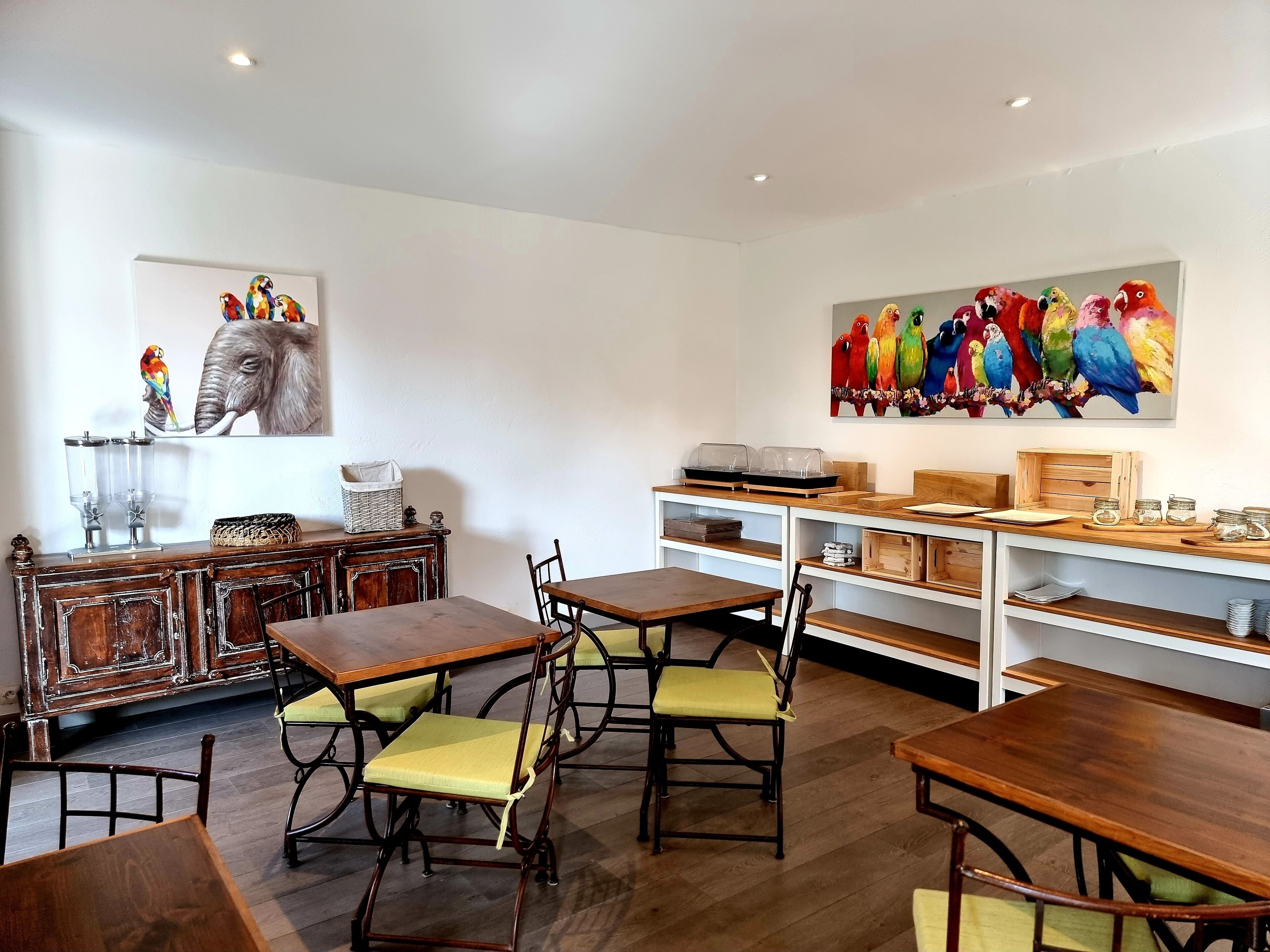 A dining area with wooden tables and chairs, a white wall with a colorful parrot painting, and a wooden cabinet with a basket.