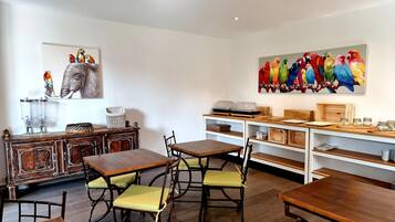A dining area with wooden tables and chairs, a white wall with a colorful parrot painting, and a wooden cabinet with a basket.