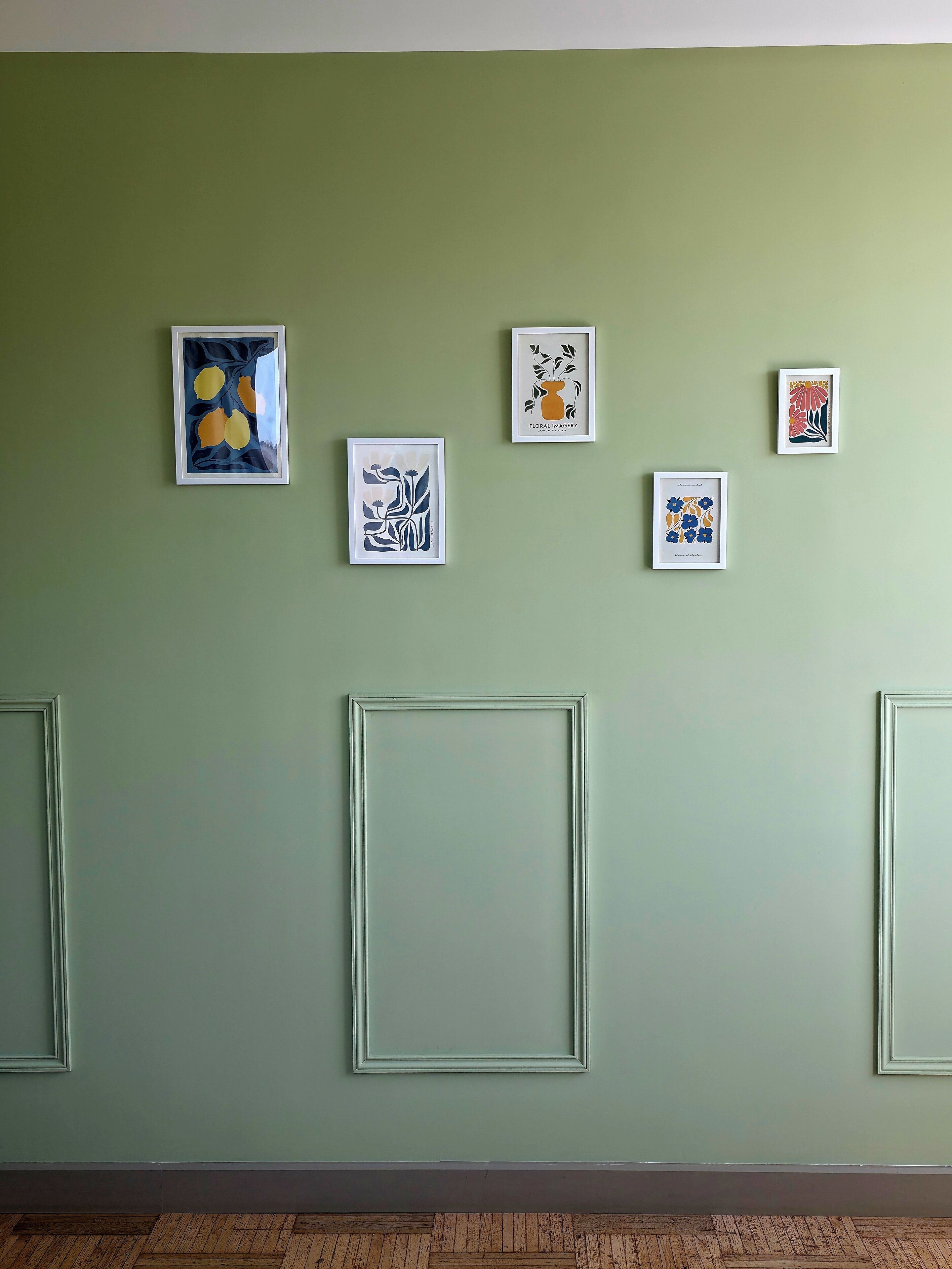 A wall with five framed artworks, a plain light green wall, and a herringbone-patterned wooden floor.