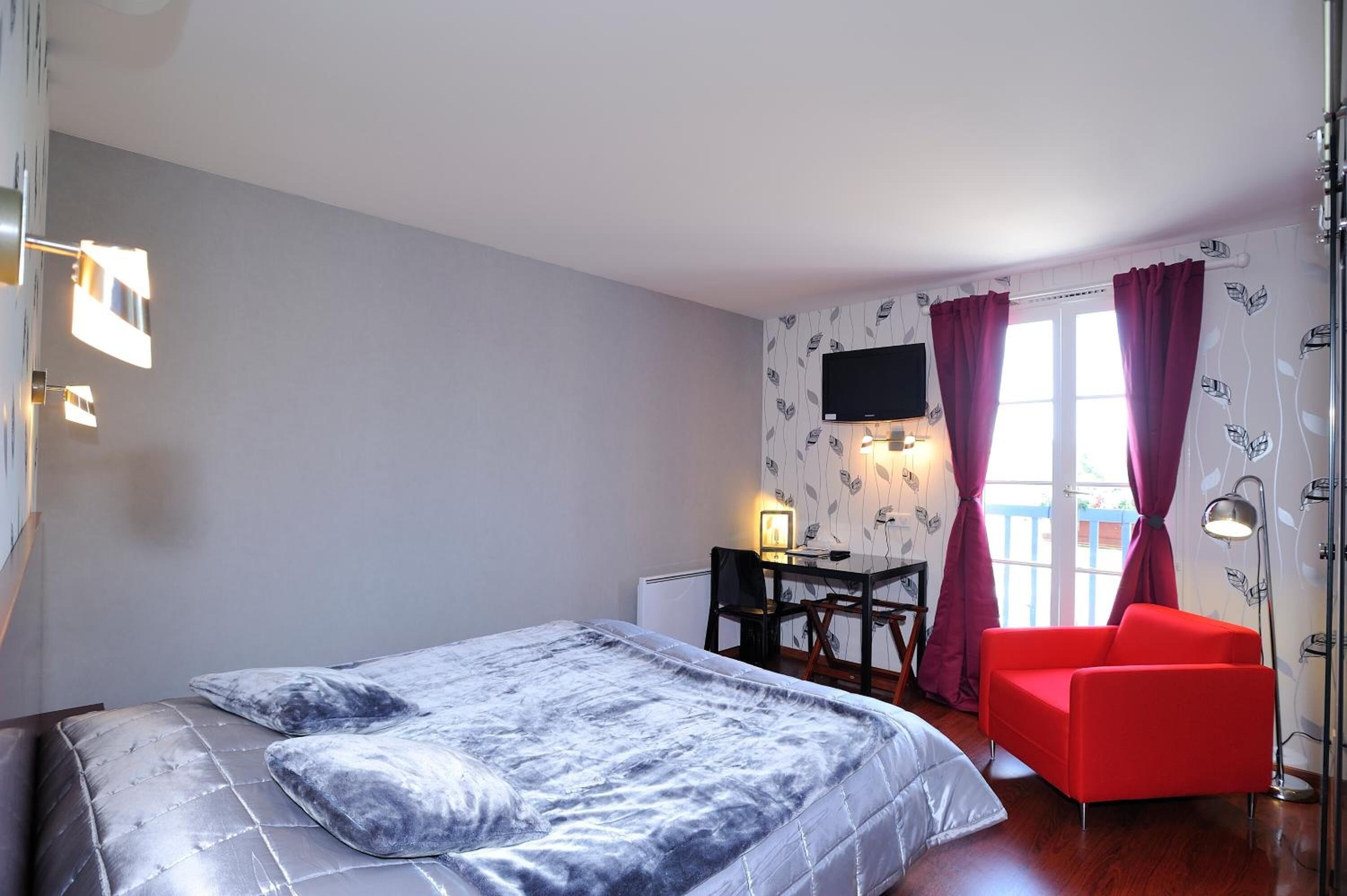 A bedroom with a bed, a desk with a chair, a red armchair, and a TV on the wall.