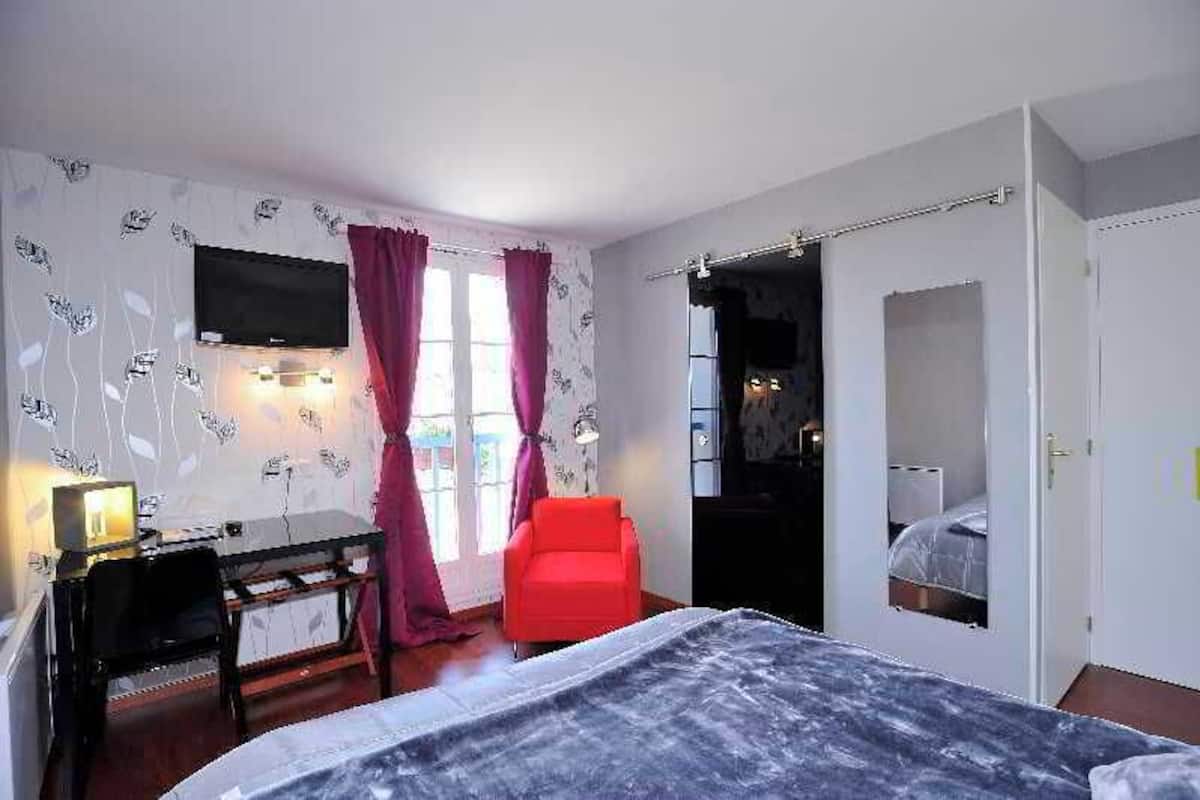 Ein Hotelzimmer mit einem Bett, einem Schreibtisch mit Lampe, einem roten Sessel, einem Fernseher und einer Tür zu einem weiteren Raum.