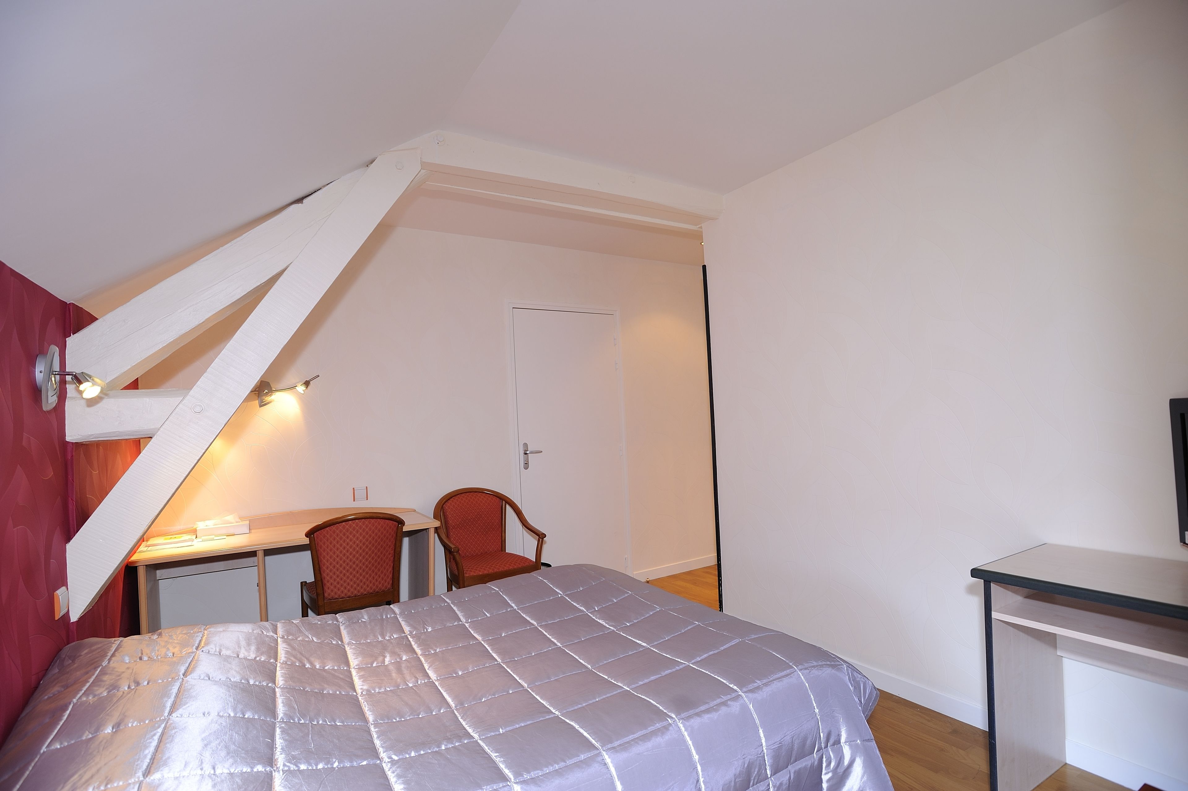 Ein Schlafzimmer mit Bett, Schreibtisch und Stuhl. Im Hintergrund sind eine Tür und eine Treppe zu sehen.