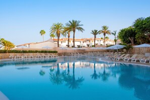 A swimming pool with lounge chairs and umbrellas, surrounded by palm trees and a building with a tiled roof.