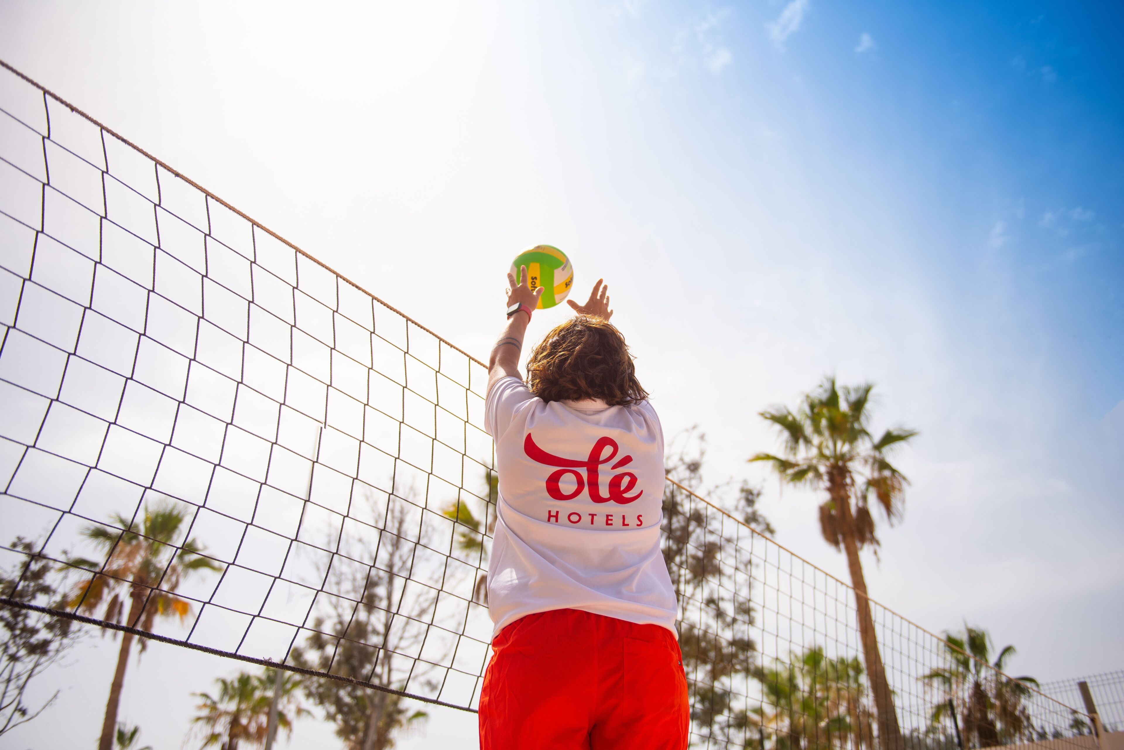 A person in a white shirt with 'ole hotels' text on the back, red shorts, and a green and yellow volleyball.