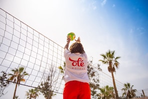 Une personne portant une chemise blanche avec lâinscription « ole hotels » dans le dos, un short rouge et un ballon de volley-ball aux couleurs verte et jaune.