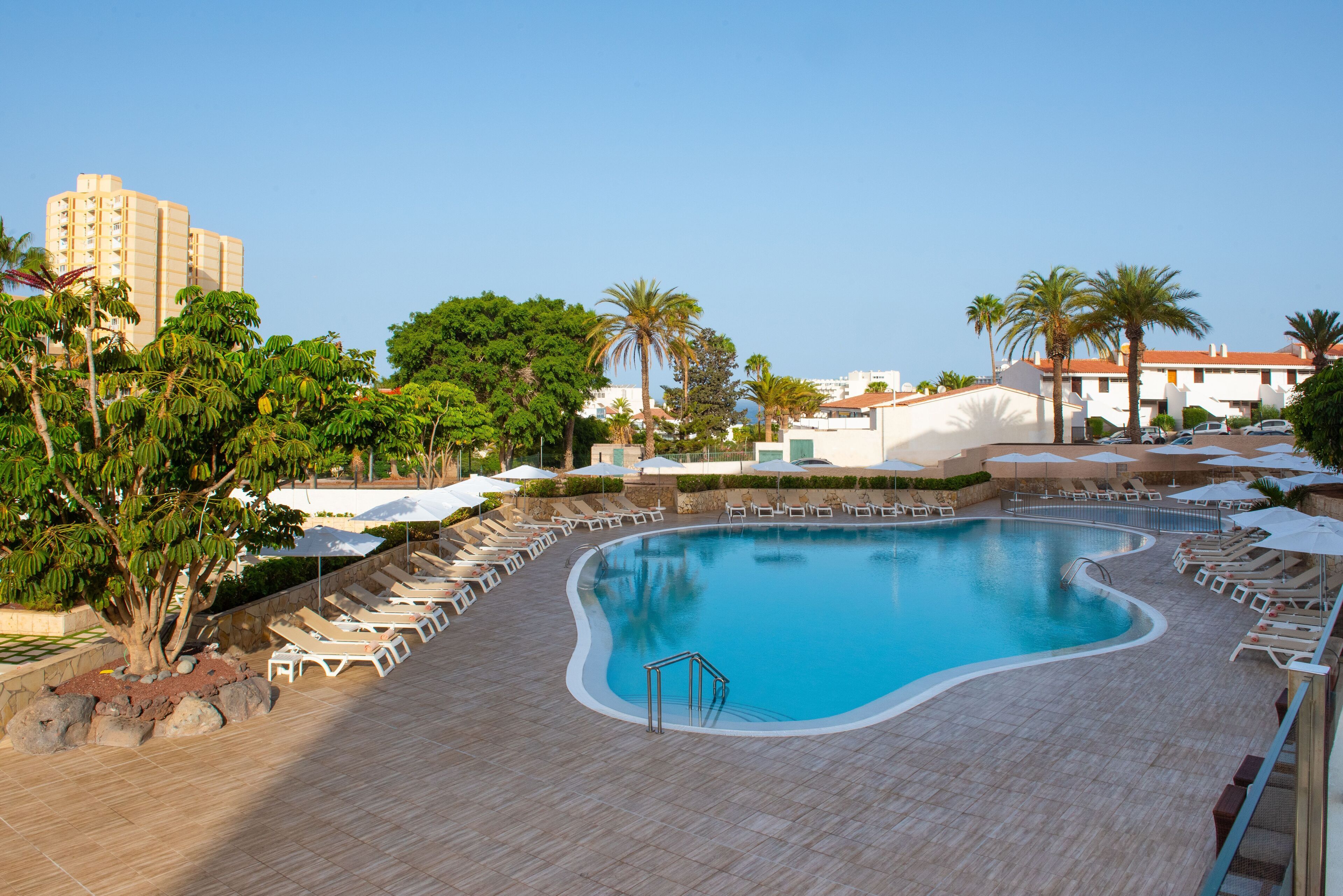 Una gran piscina con sillas de descanso y sombrillas, rodeada de palmeras y un edificio al fondo.