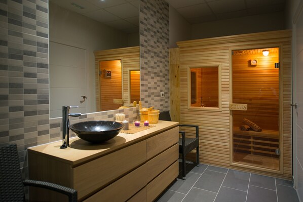 A modern bathroom with a wooden vanity, a large basin, a mirror, and a sauna visible through a glass door.