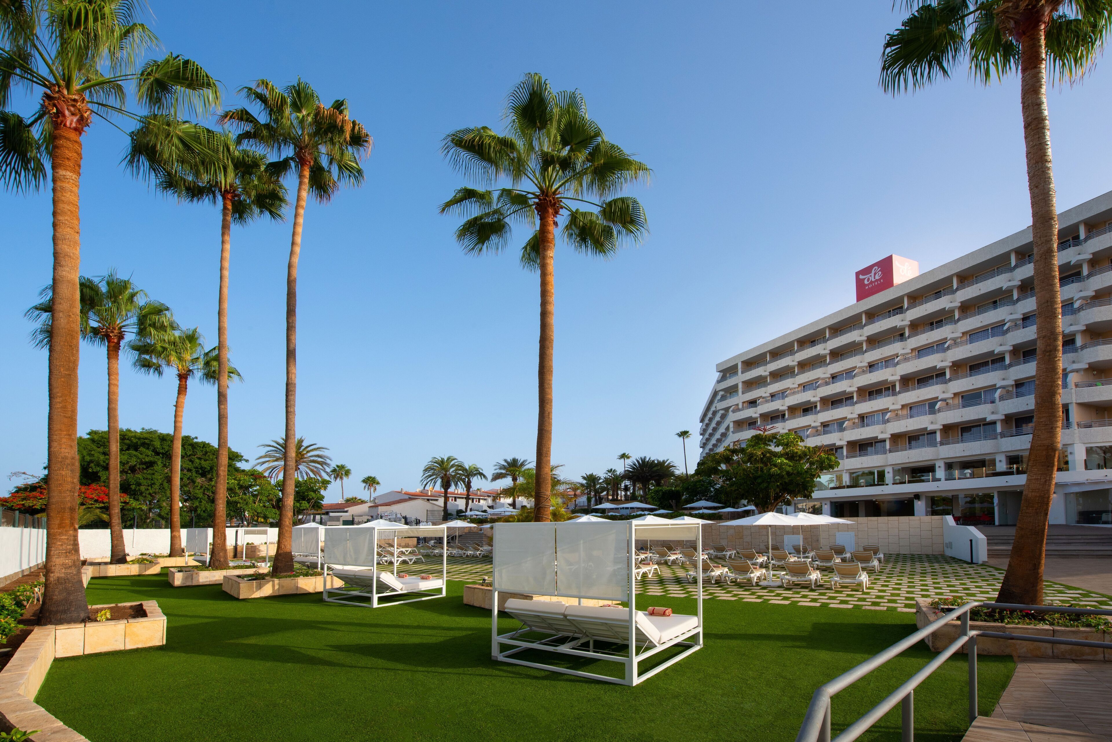 Une cour d’hôtel avec une pelouse artificielle, des palmiers, des chaises longues et un bâtiment blanc doté de balcons.