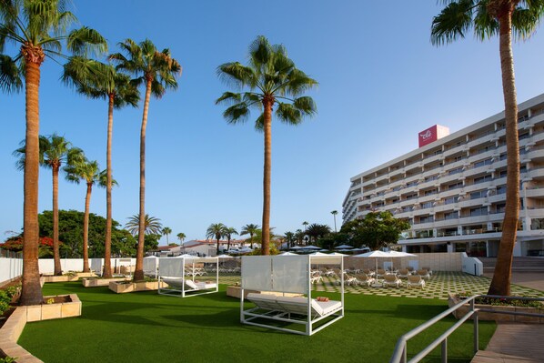 A hotel courtyard with artificial grass, palm trees, sun loungers, and a white building with balconies.