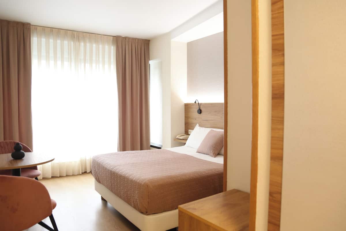 Habitación de hotel con cama, mesita de noche, una silla, escritorio y ventana con cortinas.