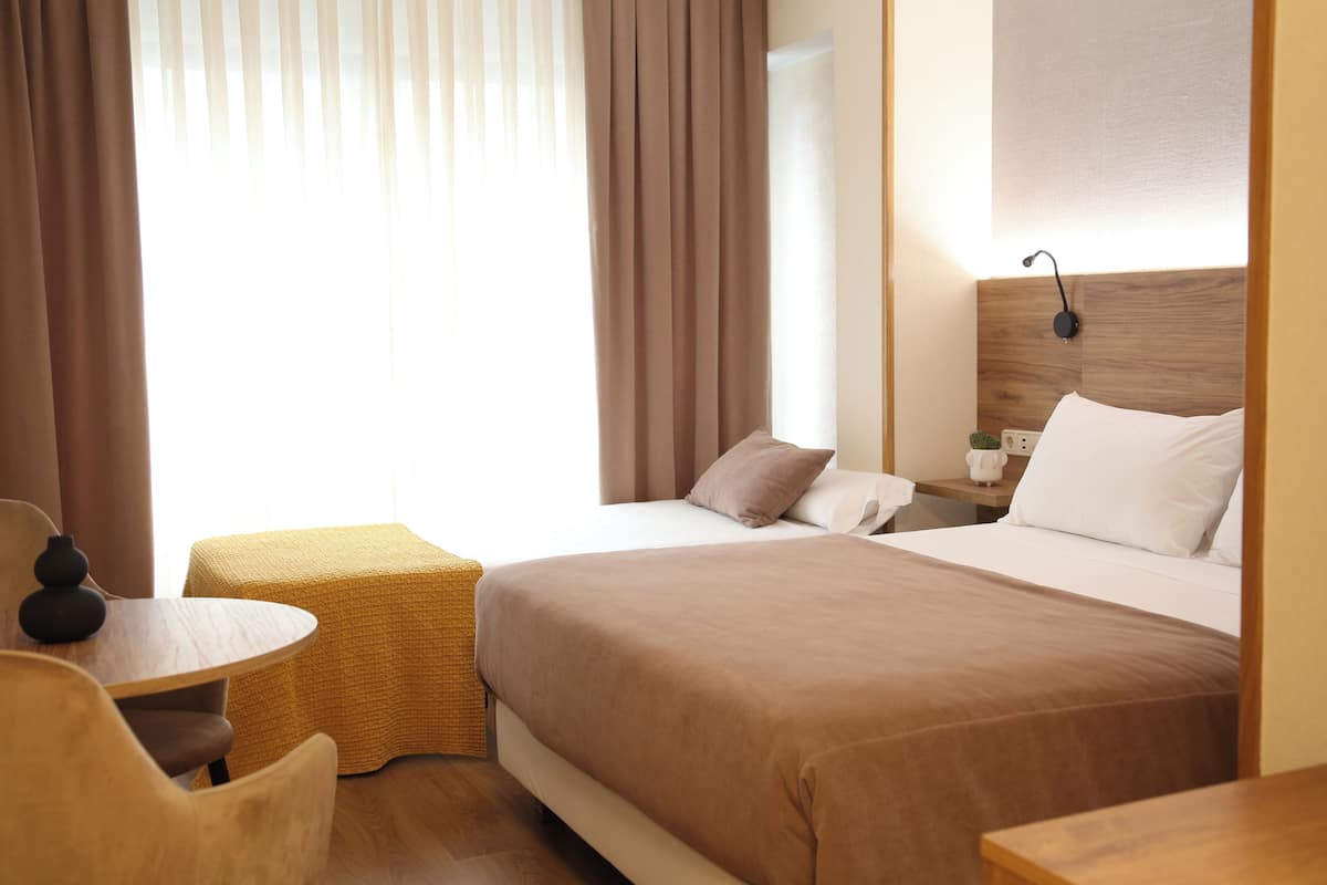 Una habitación de hotel con una cama grande, una mesita pequeña, una silla y una mesita de noche con lámpara.