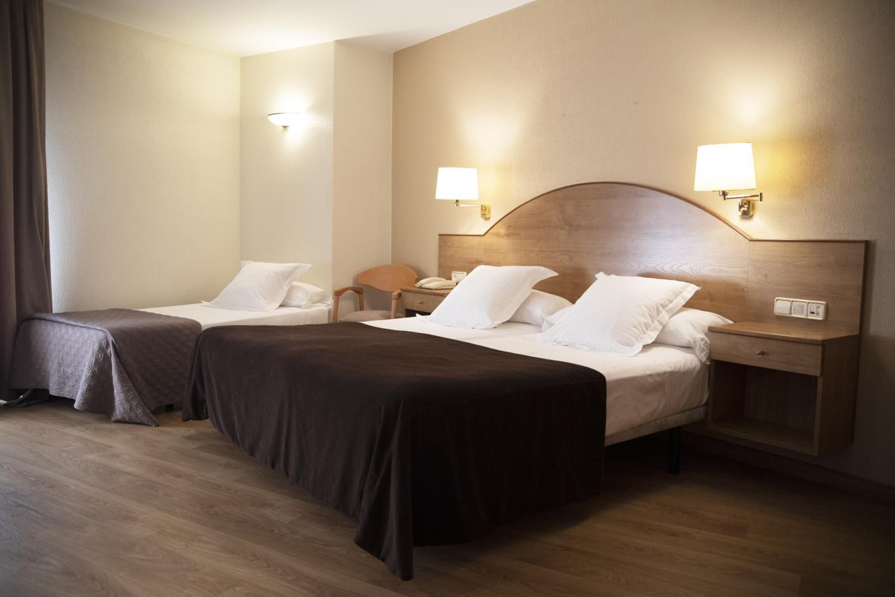 A hotel room with two beds, each with a headboard, bedside tables, lamps, and a wooden floor.