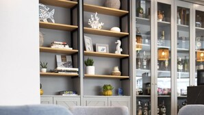 A modern living room with two grey armchairs, a round glass table, and a built-in shelving unit with decorative items and books.