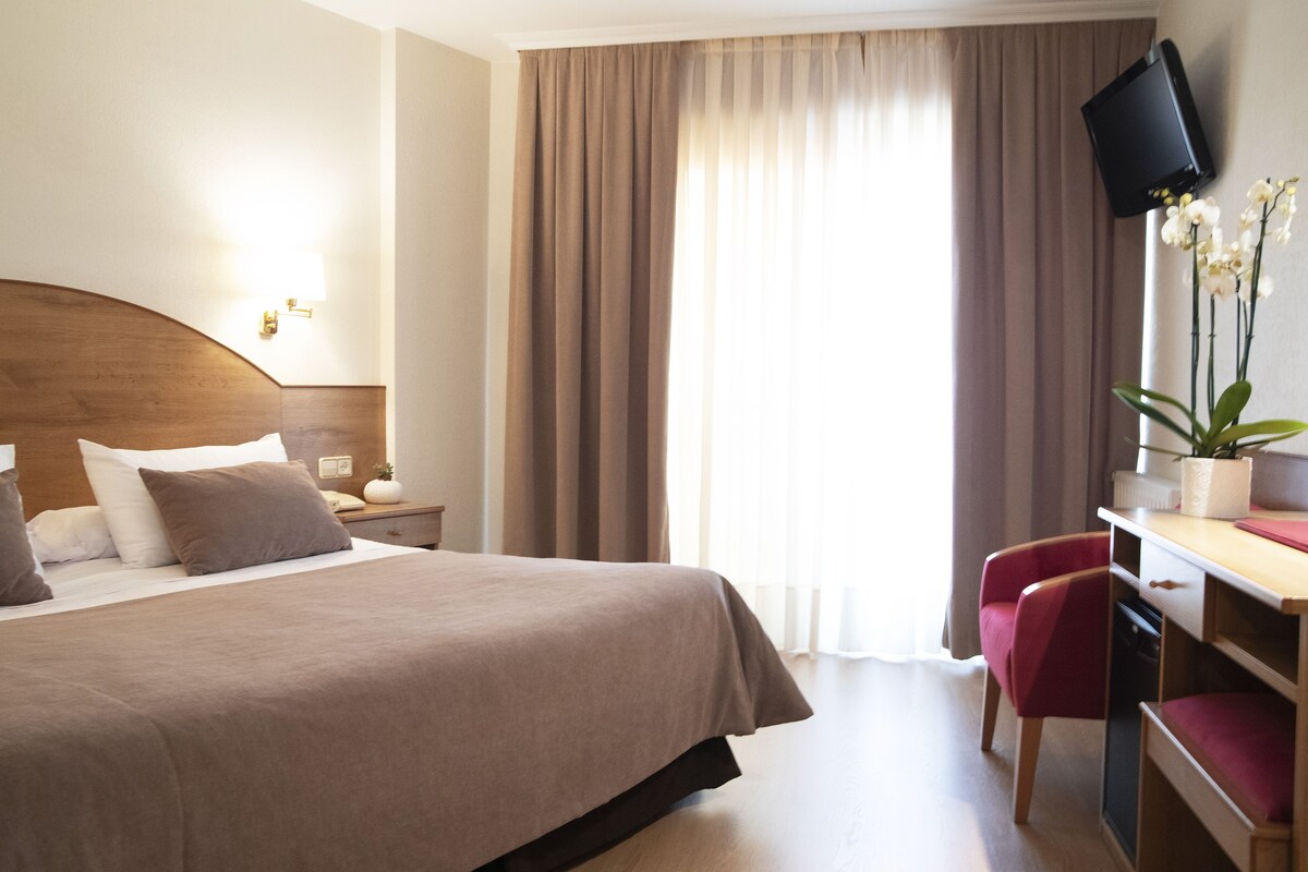 A hotel room with a large bed, a desk with a chair, a television, and a window with curtains.