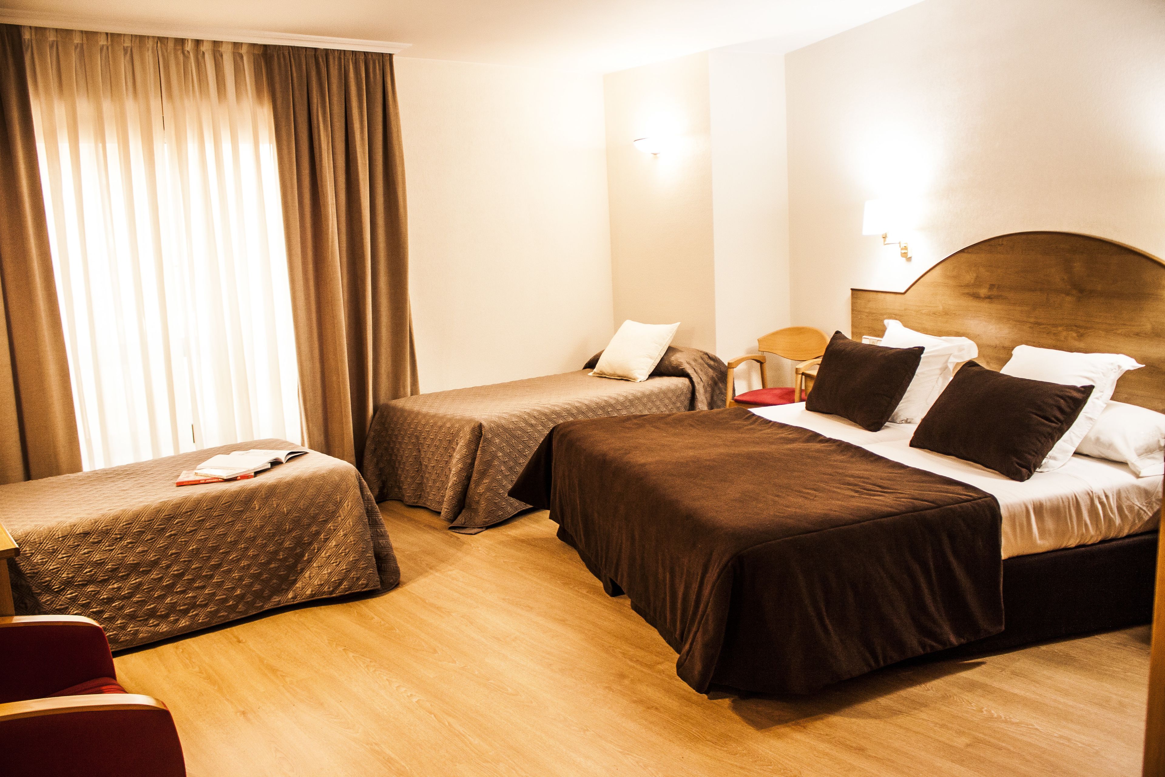 Habitación de hotel con dos camas individuales, piso de madera, un ventanal grande con cortinas y una mesita de noche con lámpara.