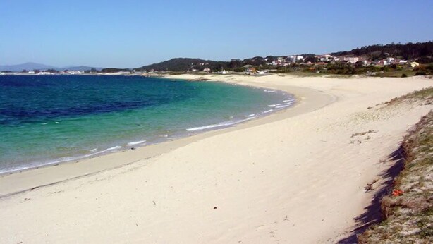 A wide sandy beach with clear blue water and a few houses in the background.
