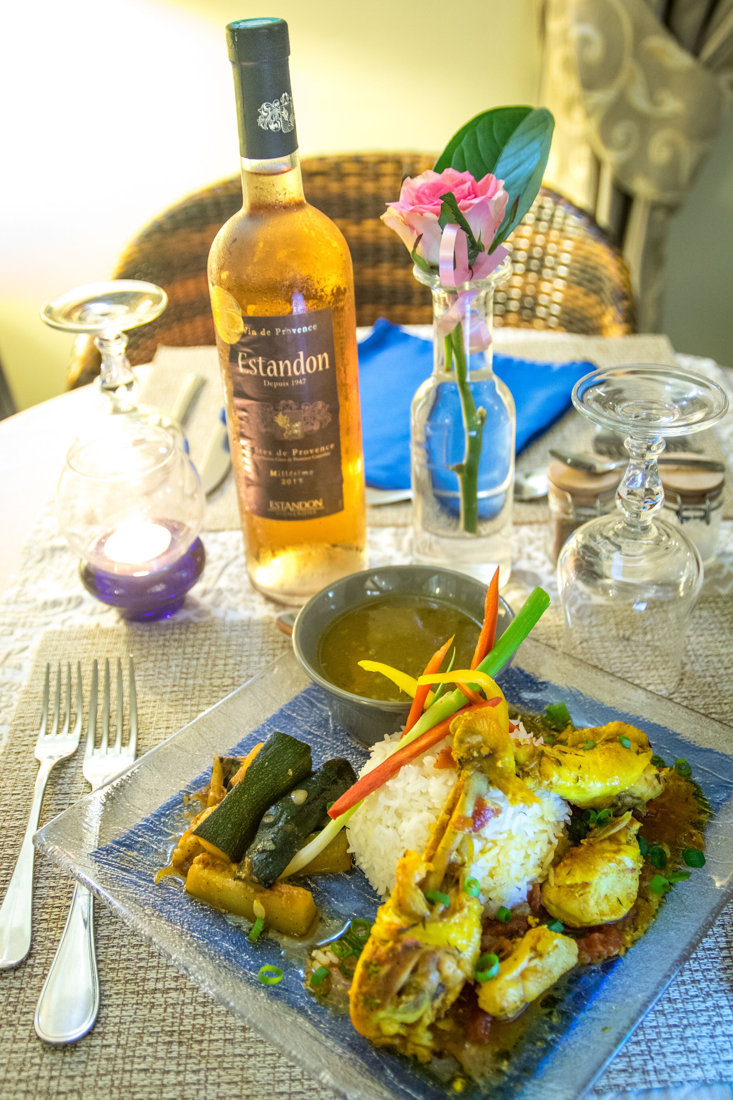 Uma mesa de jantar com uma garrafa de vinho Estandon, um copo de água, um pequeno vaso com uma flor rosa e um prato de comida.