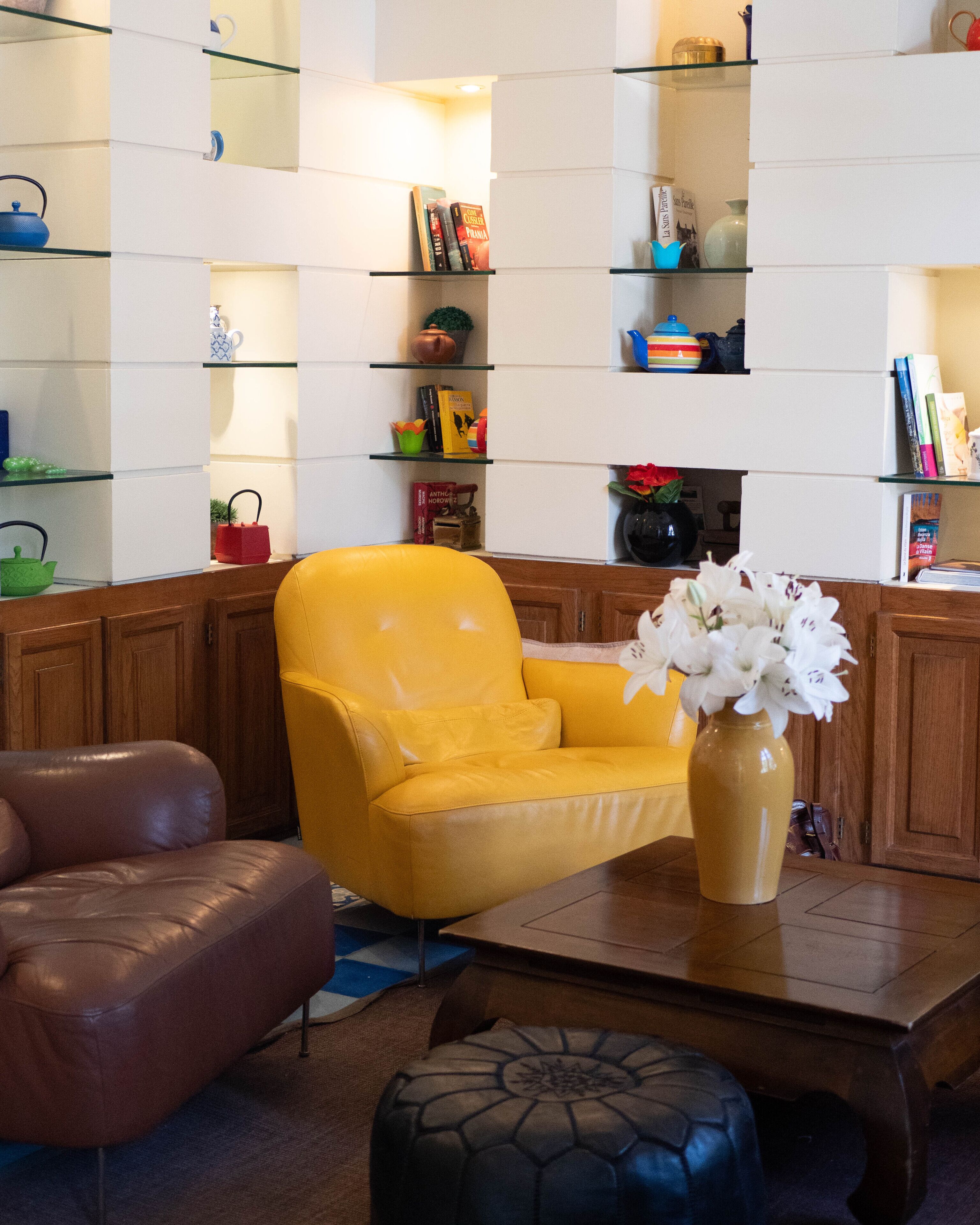 Een woonkamer met een gele fauteuil, een bruine leren bank, een salontafel en een boekenkast met diverse objecten.
