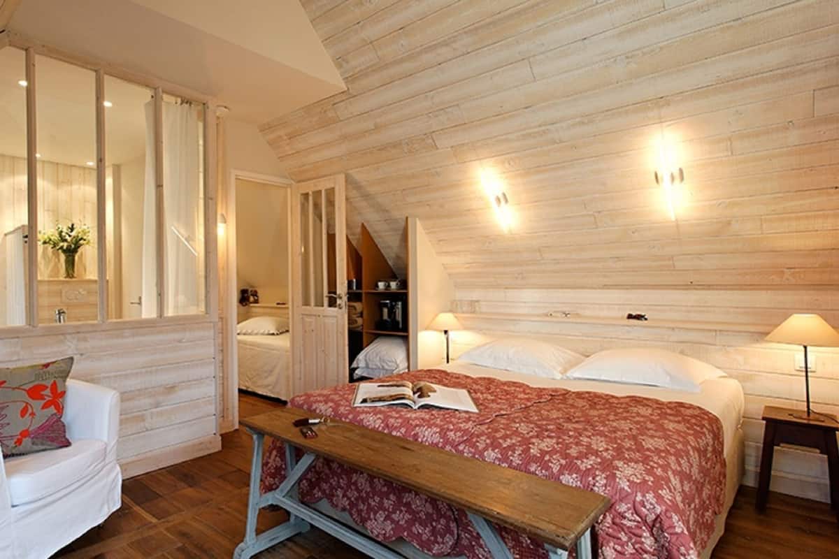 Um quarto aconchegante com teto de madeira, uma cama grande com colcha floral vermelha, uma poltrona branca e um banco de madeira.