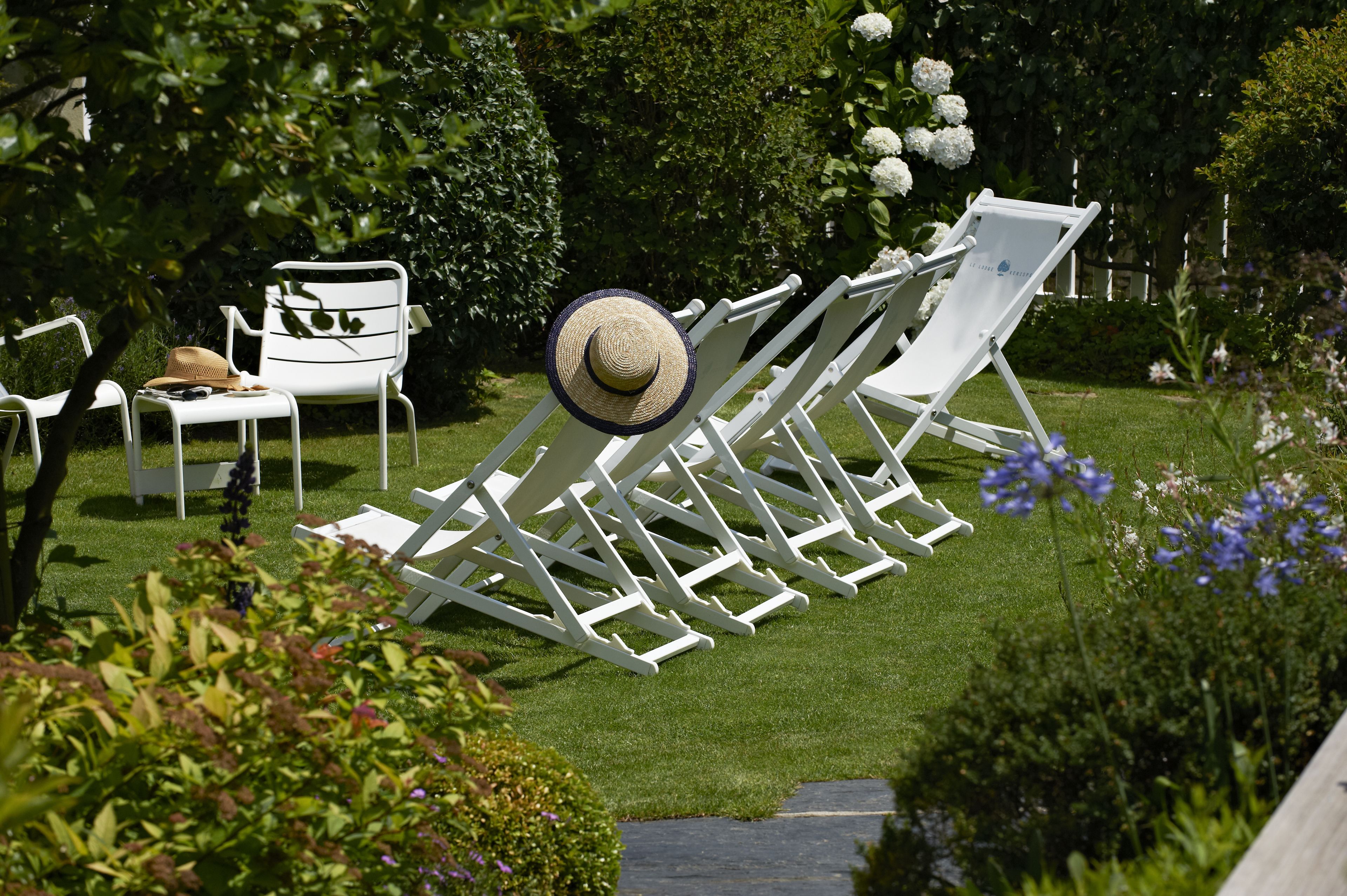 A garden with white lounge chairs and a hat on one of them.