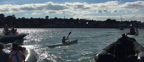 A body of water with several boats and kayaks, a distant shoreline with buildings, and a partly cloudy sky.