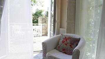 A white armchair with a floral pillow in a room with a glass door leading to an outdoor area with a stone wall and white furniture.