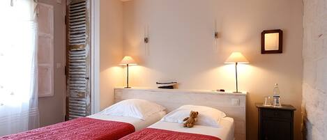 Een kamer met twee bedden, elk met een rode sprei, een nachtkastje met een lamp, en een wandplank met een boek en een fles.