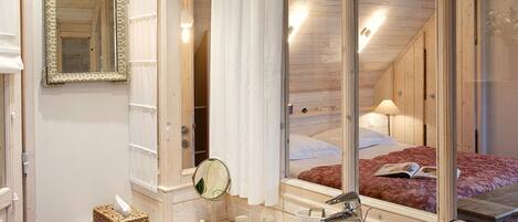 Une salle de bain avec un grand miroir, un lavabo et une fenêtre donnant sur une chambre à coucher équipée d’un lit et d’une lampe.