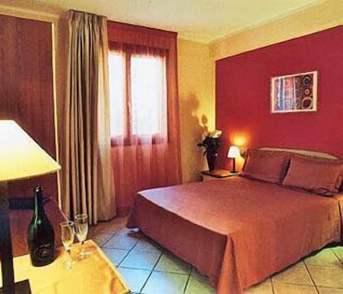 Habitació d'hotel amb un llit, una tauleta de nit amb una làmpada, una ampolla i gots sobre una taula, i una finestra amb cortines.
