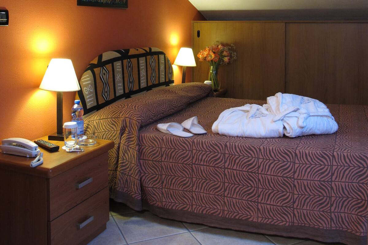 Habitación de hotel con una cama, dos mesitas de noche, una lámpara, un florero con flores y un cuadro en la pared.
