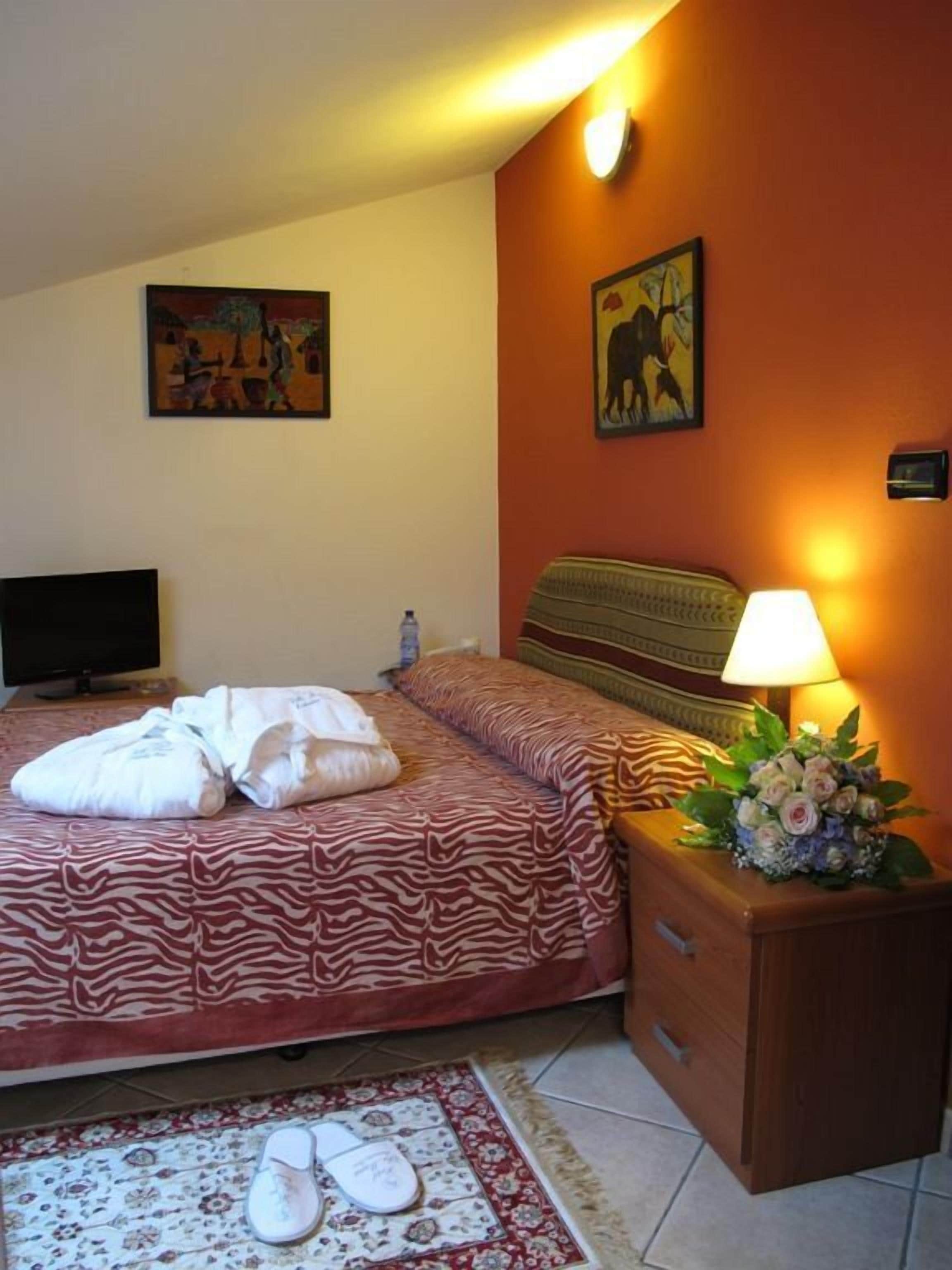 A hotel room with a bed, a TV, a nightstand with a lamp and flowers, and two paintings on the wall.