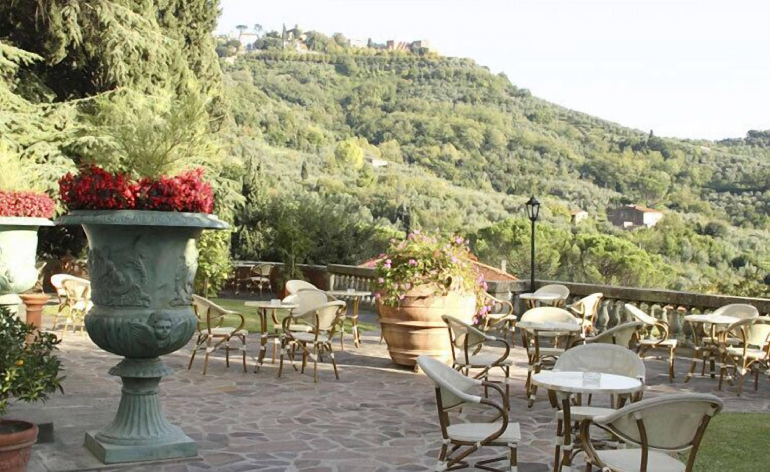 An outdoor patio with stone flooring, wicker chairs, and tables, overlooking a hillside with dense greenery.