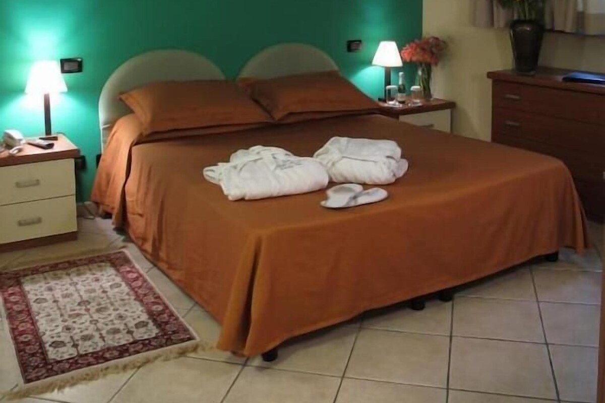 A hotel room with a bed, a nightstand, a lamp, a vase with flowers, and a painting on the wall.