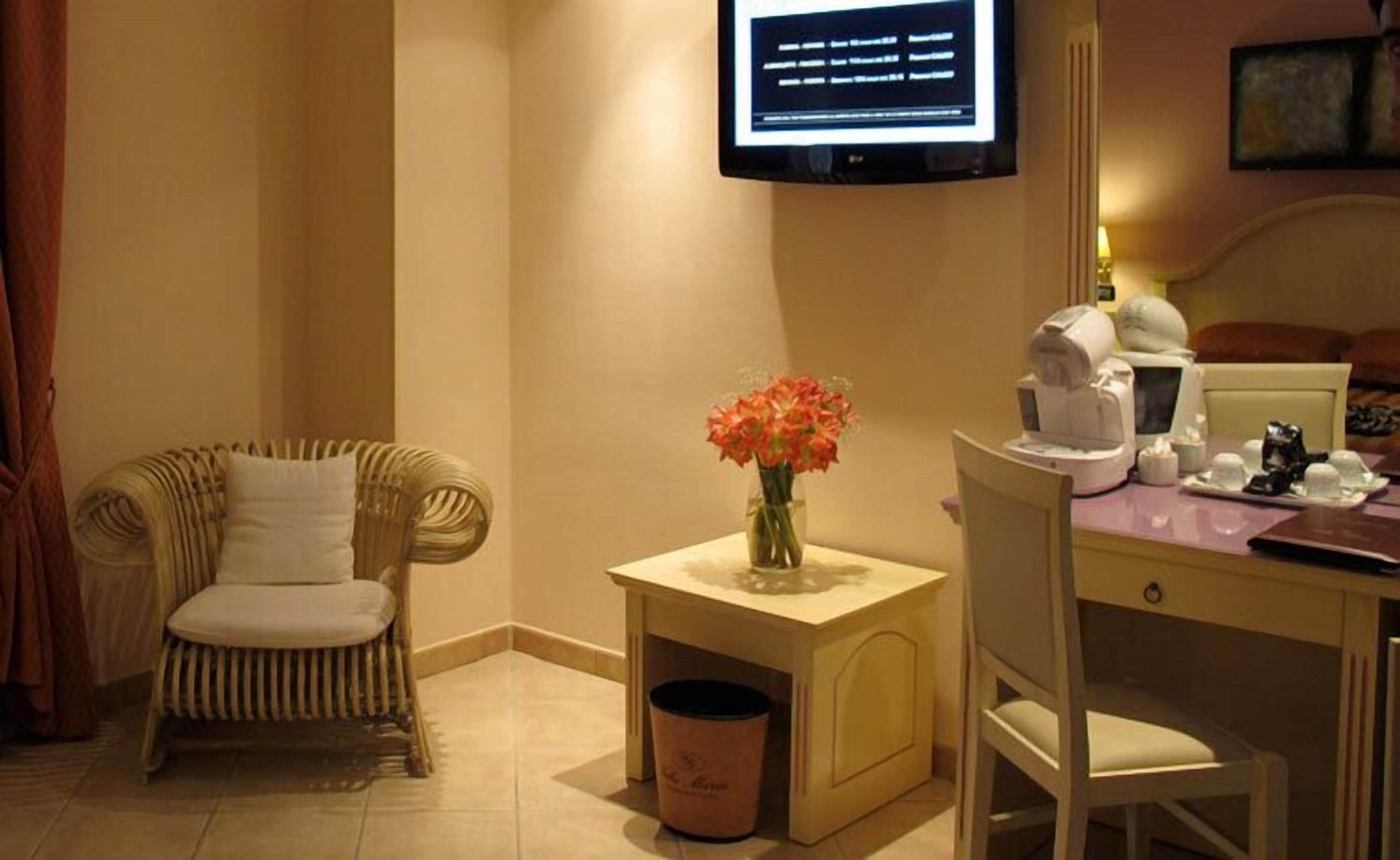 A hotel room with a bed, a chair, a small table with a vase of flowers, a coffee machine, and a TV mounted on the wall.
