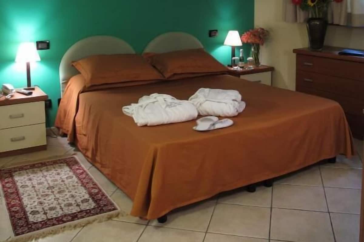 Un cuarto de hotel con una cama, una mesita de noche, una lámpara, un florero con flores y un cuadro en la pared.